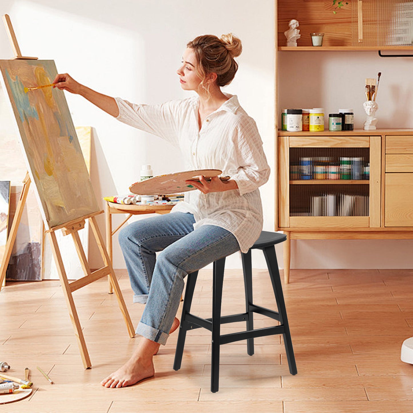Woman painting on an easel in a home studio with art supplies around.