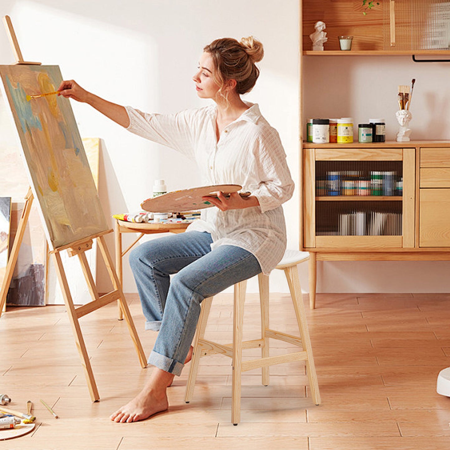 Woman painting on an easel in a home studio with art supplies around.