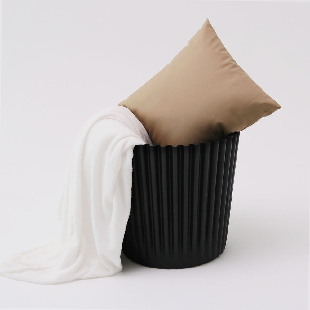 Black ribbed basket with a beige pillow and white blanket on a light gray background