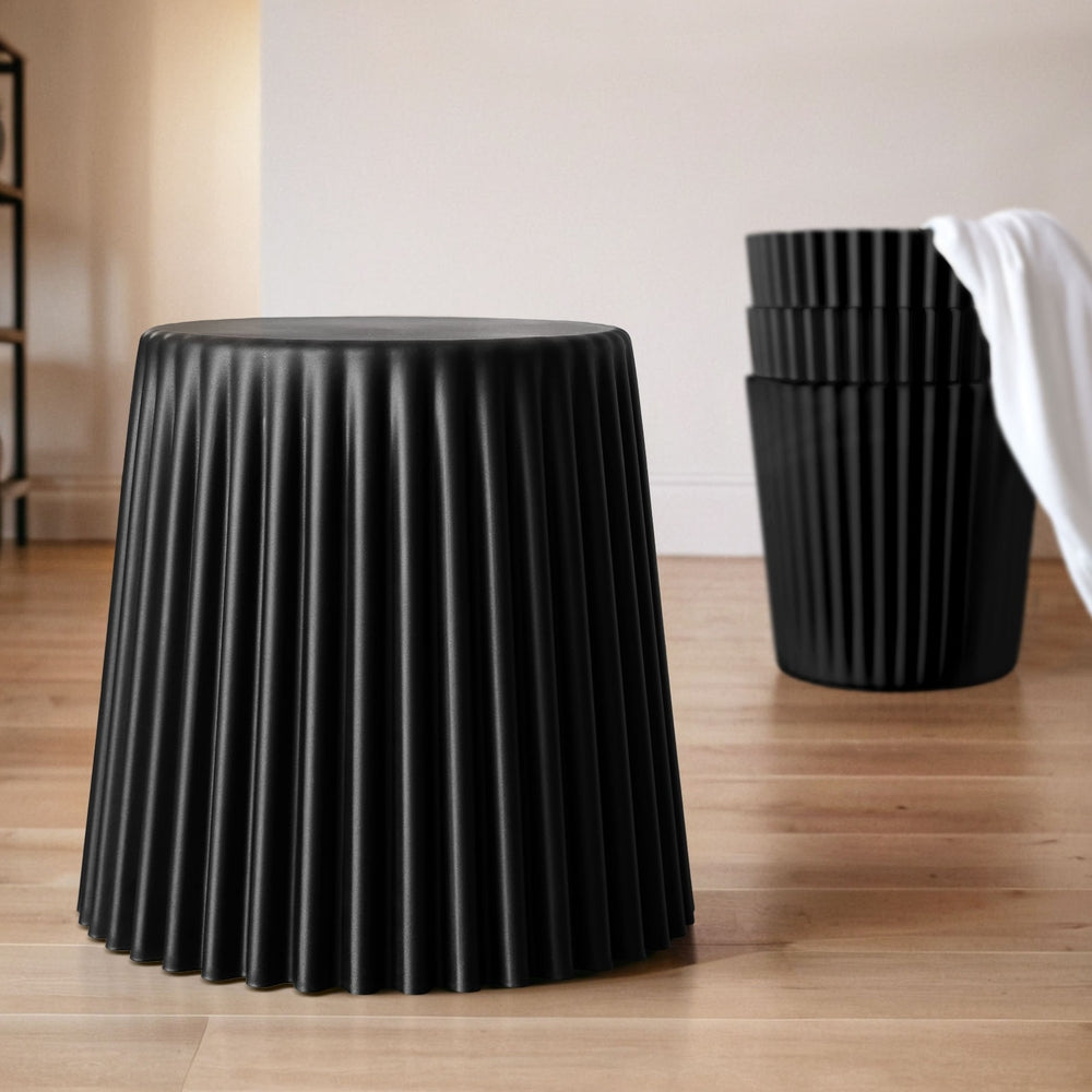 Black pleated stool on a wooden floor with a white wall in the background