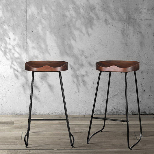 Two wooden tractor seat bar stools with metal legs against a concrete wall.