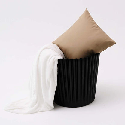 Black ribbed laundry basket with a beige pillow and white garment on a light gray background